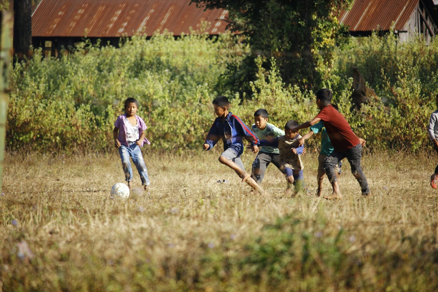 Kinder spielen Fußball auf einer Wiese mit rustikalen Gebäuden.