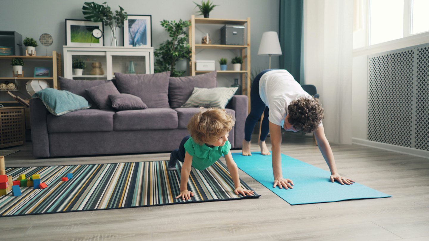 Elternteil und Kind üben Yoga-Posen auf dem Wohnzimmerboden.
